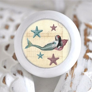 Könnte beinhalten: Weißer Keramik-Knopf mit einem runden, bunten Bild einer Meerjungfrau mit einer Muschel, umgeben von Seesternen.