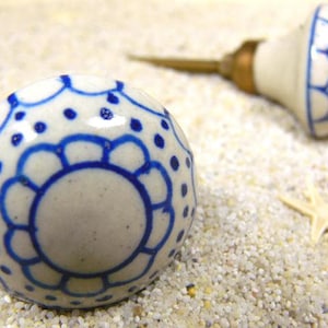 Könnte beinhalten: Zwei weiße Keramik-Türknäufe mit blauen Blumenmustern. Die Knäufe haben eine glänzende Oberfläche und liegen auf einer sandigen Oberfläche.