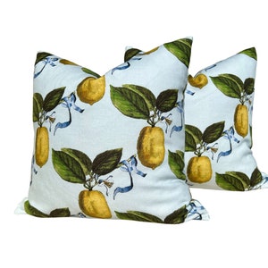 May include: Two square throw pillows with a light blue background. The pillows feature a repeating pattern of yellow lemons with green leaves and blue ribbons. The lemons are detailed with shading, giving them a realistic appearance.