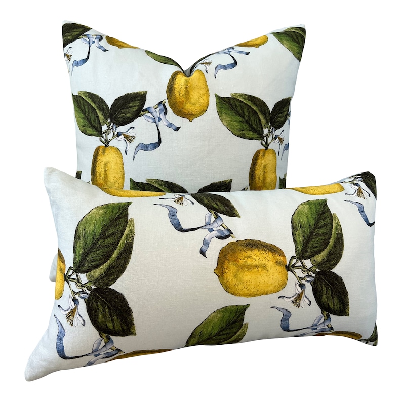 May include: Two decorative throw pillows with a white background and a repeating pattern of yellow lemons, green leaves, and blue ribbons. One pillow is rectangular, and the other is square.