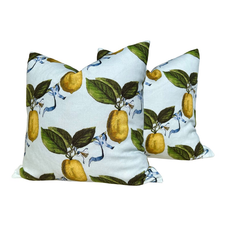 May include: Two square throw pillows with a light blue background. The pillows feature a repeating pattern of yellow lemons with green leaves and blue ribbons. The lemons are detailed with shading, giving them a realistic appearance.
