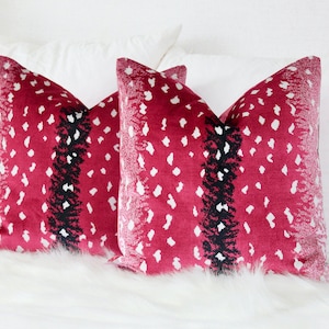 May include: Two red throw pillows with a white and black geometric pattern. The pillows are on a white bed with a white fur throw.
