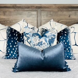 Pu&ograve; includere: Un letto con una testiera in legno e un copripiumino bianco. Ci sono diversi cuscini sul letto, tra cui due cuscini bianchi con disegni astratti blu, due cuscini in velluto blu con pois bianchi e un cuscino in raso blu tinta unita.