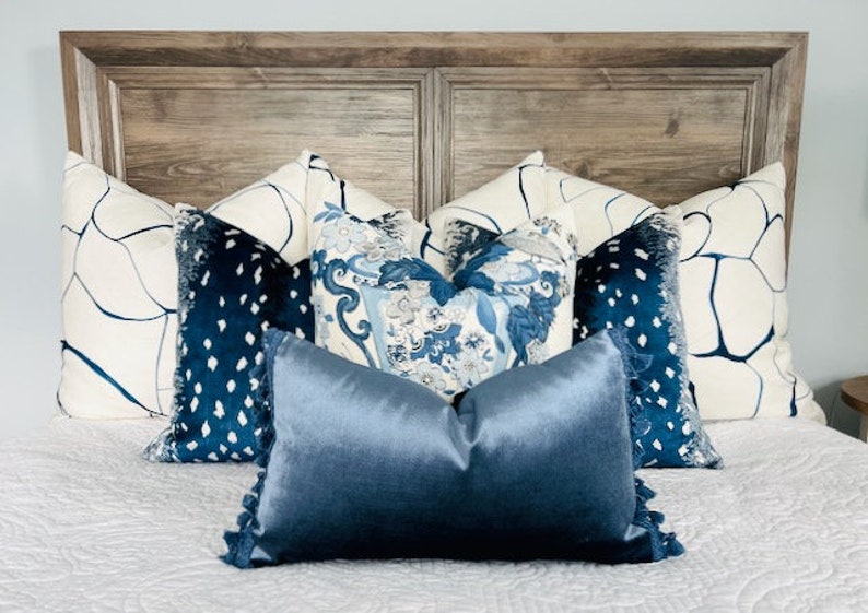Pu&ograve; includere: Un letto con una testiera in legno e un copripiumino bianco. Ci sono diversi cuscini sul letto, tra cui due cuscini bianchi con disegni astratti blu, due cuscini in velluto blu con pois bianchi e un cuscino in raso blu tinta unita.