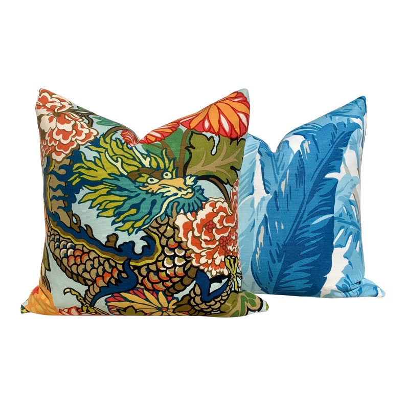 May include: Two decorative throw pillows with colorful patterns. One pillow features a blue background with a large green dragon and floral accents. The other pillow has a blue background with large white and blue leaves.
