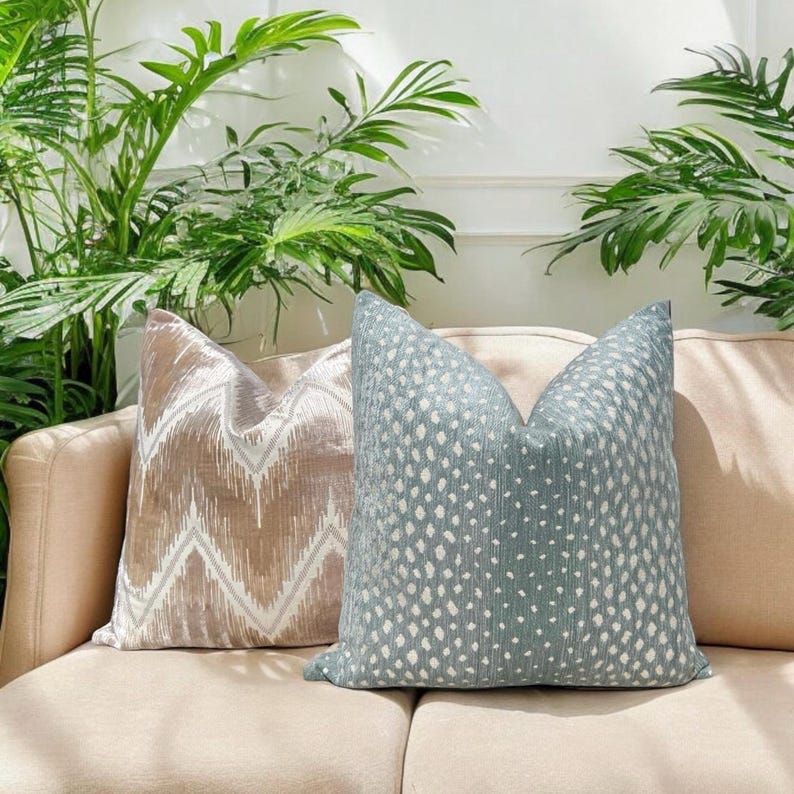 May include: Two decorative throw pillows on a beige couch. The pillow on the left is a light brown with a white chevron pattern. The pillow on the right is a light blue with a white dot pattern.