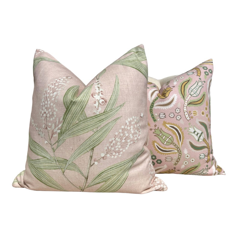 Op de afbeelding: Twee decoratieve kussens. E&eacute;n kussen heeft een lichtroze achtergrond met een botanisch ontwerp van groene bladeren en witte bloemen. Het andere kussen heeft een roze achtergrond met een bloemenpatroon in groen, geel en wit.