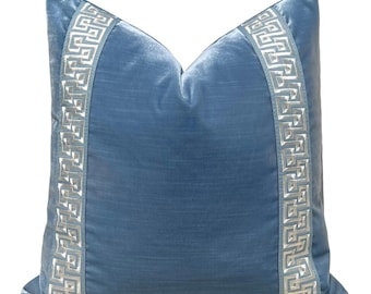 May include: A square, blue velvet pillow with a decorative border. The pillow features a Greek key pattern in a lighter shade of blue along the edges. The pillow is a soft, decorative accent for a bed or sofa.