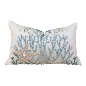 May include: Cream-colored rectangular pillow with embroidered sea life design. Features light blue coral, a beige starfish, and seahorse accents. The pillow is a decorative item for a coastal or nautical themed home.