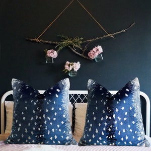 Pu&ograve; includere: Due cuscini da lancio blu e bianchi con un arrangiamento floreale bianco e rosa appeso a una parete blu scuro dietro i cuscini.