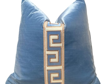 May include: A light blue velvet pillow with a decorative Greek key design in white and gold. The pillow is square-shaped and has a soft, plush appearance. The Greek key design runs vertically down the center of the pillow.