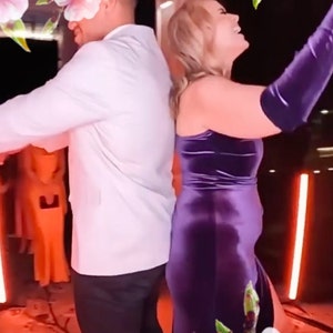 K&ouml;nnte beinhalten: Ein Paar tanzt in einem hell beleuchteten Raum zusammen. Die Frau tr&auml;gt ein violettes Samtkleid und der Mann tr&auml;gt ein wei&szlig;es Hemd und eine schwarze Hose. Im Hintergrund befinden sich rosa Blumen.