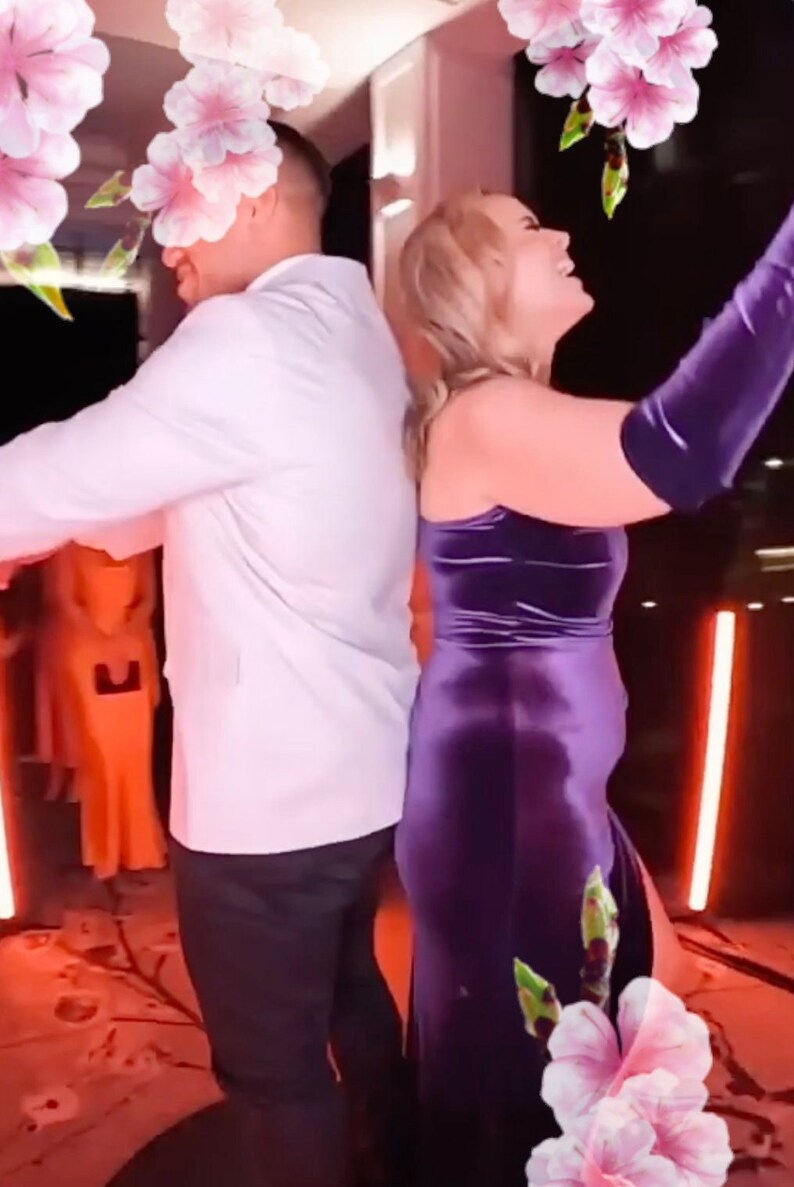 K&ouml;nnte beinhalten: Ein Paar tanzt in einem hell beleuchteten Raum zusammen. Die Frau tr&auml;gt ein violettes Samtkleid und der Mann tr&auml;gt ein wei&szlig;es Hemd und eine schwarze Hose. Im Hintergrund befinden sich rosa Blumen.