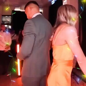 K&ouml;nnte beinhalten: Ein Mann und eine Frau tanzen in einem schwach beleuchteten Raum. Der Mann tr&auml;gt einen grauen Anzug und die Frau ein leuchtend oranges Kleid. Im Hintergrund befinden sich bunte Lichterketten.