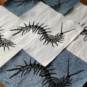 May include: A patchwork quilt square arrangement featuring alternating light cream and blue denim fabric squares. Each square displays a black ink-style print of a segmented centipede with multiple legs and long antennae, finished with pinked edges.