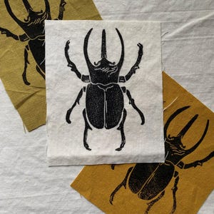 May include: Three fabric squares with a black beetle design. The central square is white, while the other two are mustard yellow. The beetle illustration is detailed, with prominent horns and legs. The fabric has a textured appearance.