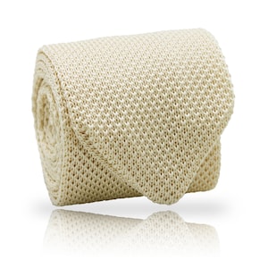 May include: A rolled-up, cream-colored knitted tie. The tie is made of a textured fabric with a visible weave pattern. The tie is folded over itself, with the pointed end visible. The background is white, and the tie casts a shadow.