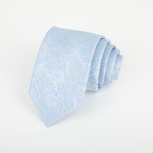 May include: A light blue necktie featuring a tonal floral pattern with leaves and blossoms woven into the fabric. The tie is rolled up, displaying a textured weave and a pointed tip, showcasing a consistent pale blue colour throughout the design.
