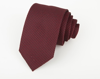 May include: A deep burgundy necktie featuring a fine, textured knit pattern. The tie is rolled into a coil, showcasing the consistent woven fabric structure and the rich, dark red colour throughout the entire length of the accessory.