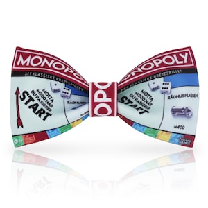 May include: A novelty bow tie featuring a Monopoly board game design. The bow tie has a light blue background with game board elements, dice, and the word "START". The text "MONOPOLY" is displayed in red on the top edge.