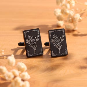 May include: A pair of black cufflinks with a floral design. Each cufflink has a rectangular shape with a silver floral illustration and a decorative border. The cufflinks are set against a wooden surface with soft, blurred floral accents.