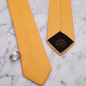 May include: A solid orange necktie with a smooth finish and a silver-toned metal ring. The back of the tie features a black fabric lining with a detailed embroidered graphic of a dog face, showcasing a textured appearance and a pointed tip.