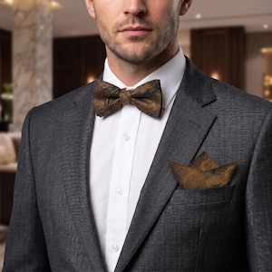 May include: A formal ensemble featuring a patterned brown bow tie and matching pocket square. The bow tie and pocket square have a detailed design. The outfit includes a gray suit jacket and a white dress shirt.