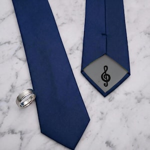 May include: A solid navy blue necktie with a smooth finish and a silver-toned metal ring. The underside of the tie features a grey fabric lining with a black treble clef musical symbol printed in the center of the pointed tip.