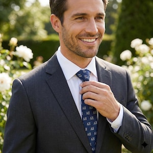 Può includere: Un abito grigio scuro con una camicia bianca e una cravatta blu. La cravatta presenta un motivo ripetuto delle lettere "AH". La persona sorride e si sistema la cravatta.