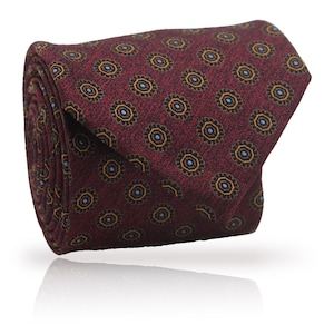 May include: Rolled burgundy tie with a repeating floral pattern in gold and blue. The tie is made of a textured fabric and is rolled up, showcasing the pattern. The floral design is evenly spaced across the tie.