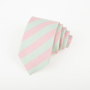 Pode incluir: Uma gravata com um padrão de riscas diagonais que alterna entre verde menta claro e rosa pastel suave. O tecido tem uma textura tecida visível em toda a superfície e a gravata está dobrada numa forma enrolada e sobreposta.