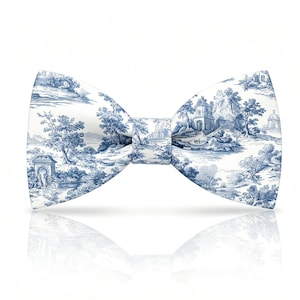 May include: A white bow tie with a blue toile pattern featuring a scenic landscape with trees, buildings, and water. The bow tie is symmetrical and reflects on the white surface below.