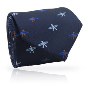 Puede incluir: Corbata enrollada de color azul marino con un patrón repetido de surfistas con tablas de surf. Los surfistas están representados en blanco, azul claro y marrón. La corbata está hecha de una tela suave y es adecuada para ocasiones formales o informales.