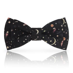 Puede incluir: Una pajarita negra con un diseño celestial de estrellas, lunas y planetas en blanco, rosa y dorado. La pajarita tiene una forma clásica, perfecta para eventos formales o para añadir un toque único a cualquier atuendo.