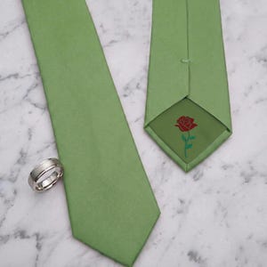May include: A sage green necktie featuring a red rose graphic printed on the interior lining of the pointed tip, accompanied by a polished silver-toned metal band ring with a brushed center finish and smooth, reflective beveled edges.