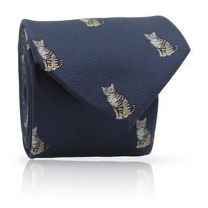 May include: Rolled navy blue tie with a repeating pattern of seated tabby cats. The cats are in shades of brown, tan, and grey. The tie is made of a smooth fabric and is neatly folded.