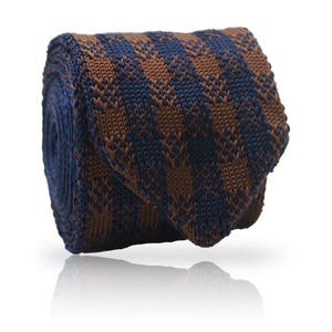 Può includere: Cravatta in maglia arrotolata con motivo a quadri blu navy e marrone. La cravatta è realizzata in un materiale strutturato e viene esposta su sfondo bianco con un riflesso. Il motivo è un design geometrico ripetitivo.