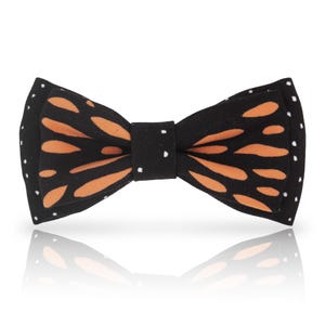May include: A black bow tie with an orange and white pattern resembling a butterfly wing. The bow tie has white polka dots on the ends and is centered on a white background.