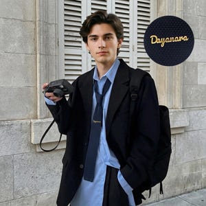 May include: A black blazer worn over a light blue button-down shirt with a dark blue knit tie featuring the gold embroidered text "Dayanara". A black camera is held in one hand, and a black backpack is worn over one shoulder against a white wall.