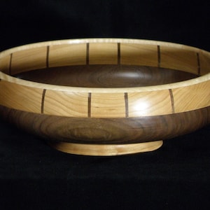 Op de afbeelding: Een handgemaakte houten kom met een lichtgekleurde rand en een donkerbruine basis. De rand heeft verticale donkerbruine inlegstukken. De kom heeft een afgeronde vorm en staat op een kleine, cirkelvormige basis. De binnenkant is donkerbruin.