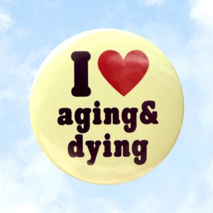 May include: A round, light yellow button with the words "I ❤️ aging & dying" in black and a red heart. The button is set against a blue sky background with white clouds. Text on the left and right sides of the button reads "pins magnets totes" and "valentinemotel.com".