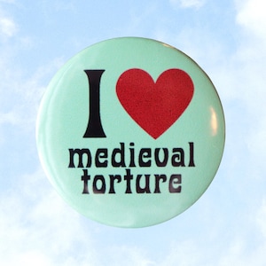 May include: A round, light green button with the words "I" and "medieval torture" in black, and a red heart. The button is set against a blue sky background with white clouds. The button has the text "valentinemotel.com" on the right side.