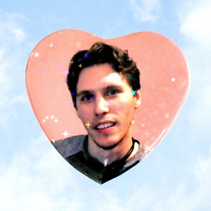 May include: A heart-shaped image featuring a person's face, set against a light blue sky with clouds. The heart is a soft pink with sparkling star accents. The text "ValentineMotel.com" is at the bottom.