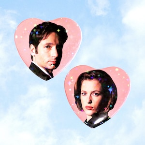May include: Two pink heart-shaped stickers with images of a man and a woman. The stickers have a holographic star pattern and are set against a blue sky background. The text "ValentineMotel.com" is at the bottom.