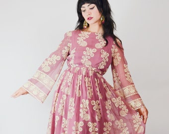 May include: A long, flowing mauve dress with long, bell sleeves. The dress features an all-over floral pattern in a cream color, with a matching cream border at the hem and sleeve cuffs. The dress has a cinched waist and a round neckline.