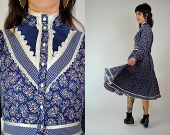 Puede incluir: Vestido vintage azul marino con estampado floral, cuello alto, mangas largas y cinturón de lazo. El vestido presenta detalles de encaje blanco y lunares. El vestido llega aproximadamente a la rodilla.