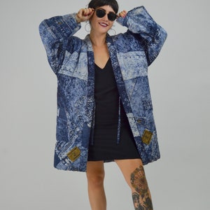 Op de afbeelding: Een blauwe spijkerjas met een patchwork design. De jas heeft een lange, losse pasvorm en heeft verschillende blauwtinten. Het heeft een kraag, zakken en een knoopsluiting. De jas wordt over een zwarte jurk gedragen.