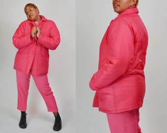 May include: A bright pink quilted jacket with a collared neckline and two front pockets. The jacket is paired with matching pink pants. The outfit is completed with black boots. The jacket has a button-up closure.