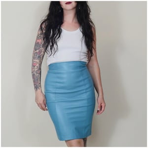 May include: A light blue leather pencil skirt. The skirt is a classic knee-length design with a high waist. The skirt is a solid colour. The model is wearing a white vest top. The skirt is approximately 60 cm in length.