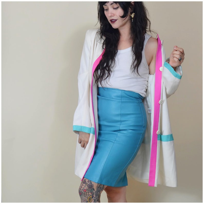 May include: A cream coat with pink and turquoise trim is worn over a turquoise pencil skirt. The coat has large white buttons and a pink stripe down the front. The skirt is a solid turquoise colour.